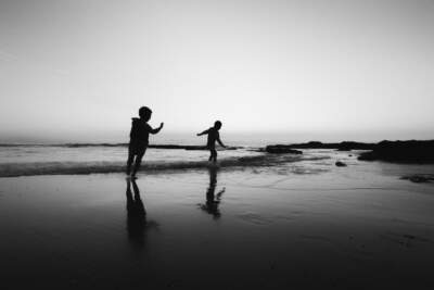 Two children playing at low tide on a beach near rocks at sunrise in black and white silhouetted. (Getty Images)